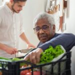 Older Black man receiving fresh vegetables from a community food program, with a younger volunteer assisting in the background—representing food security and care provided by charities in B.C