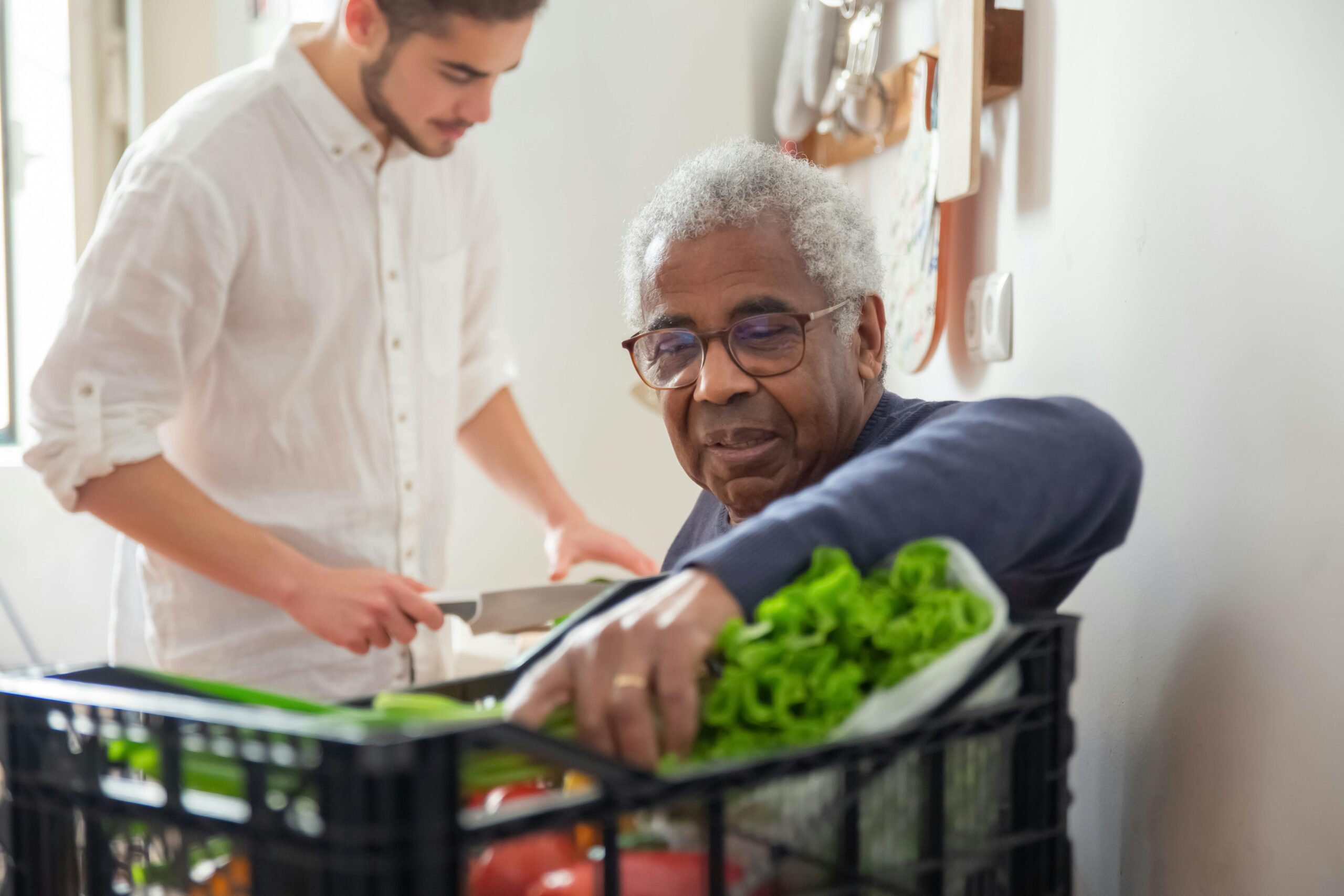 Older Black man receiving fresh vegetables from a community food program, with a younger volunteer assisting in the background—representing food security and care provided by charities in B.C