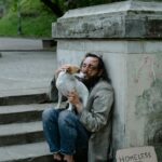 A man experiencing homelessness sits on stone steps hugging his small dog, beside a cardboard sign that reads “Homeless and Hungry.”