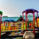 A child and an adult standing on a colorful, accessible outdoor playground structure outside a KidsAbility facility
