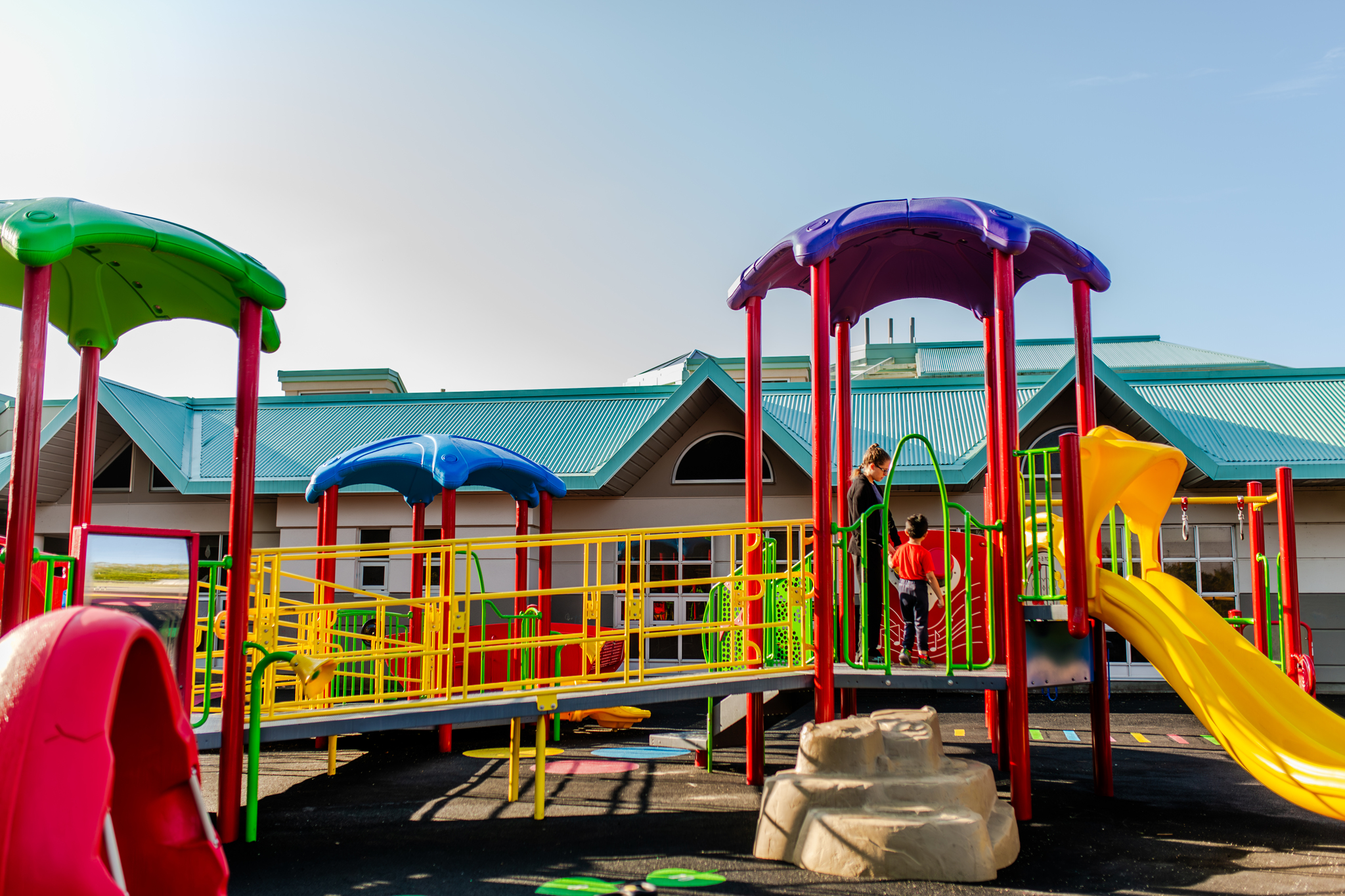 A child and an adult standing on a colorful, accessible outdoor playground structure outside a KidsAbility facility