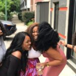 Five Black women smiling and spending time together outdoors in a bright natural setting.