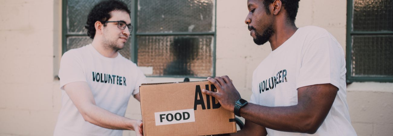 Two male volunteers wearing white volunteer shirts holding a cardboard food aid donation box outside.